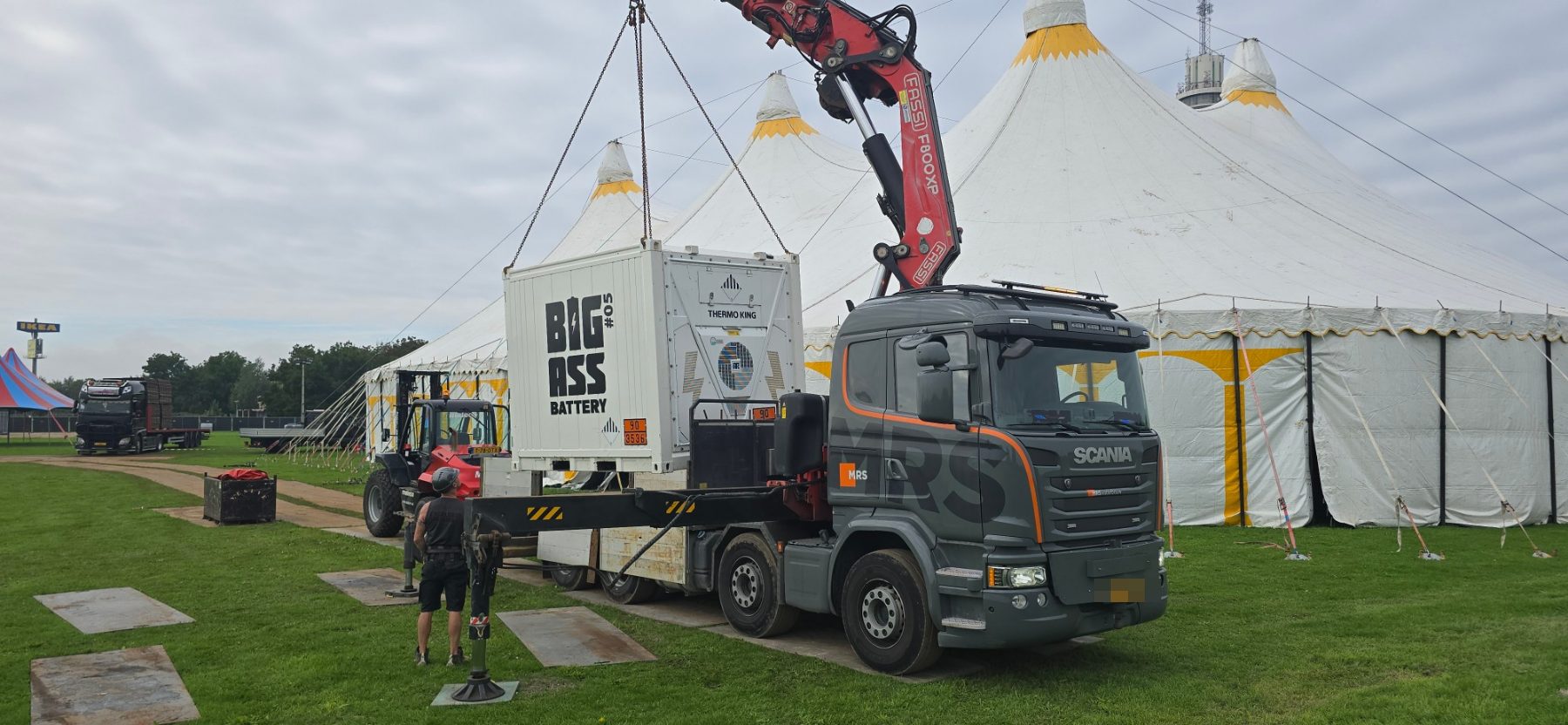 Kraanwagen tilt een accucontainer naast een festivaltent
